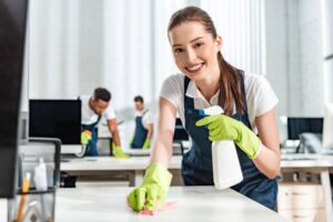 A professional office cleaner smiling and wiping a desk and holding a spray bottle with other cleaners in the background