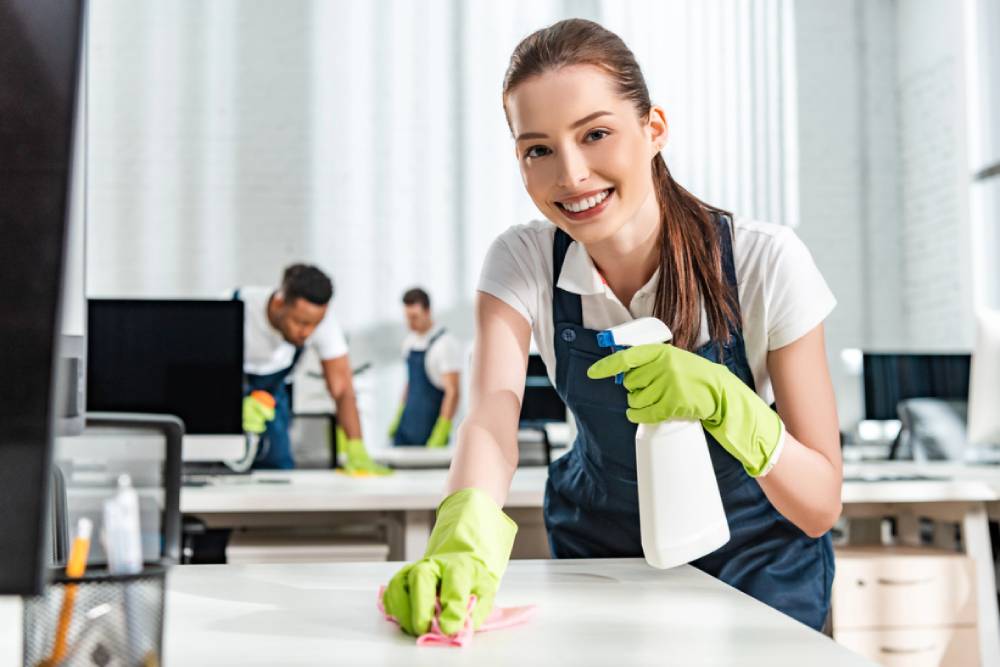 A professional office cleaner smiling and wiping a desk and holding a spray bottle with other cleaners in the background